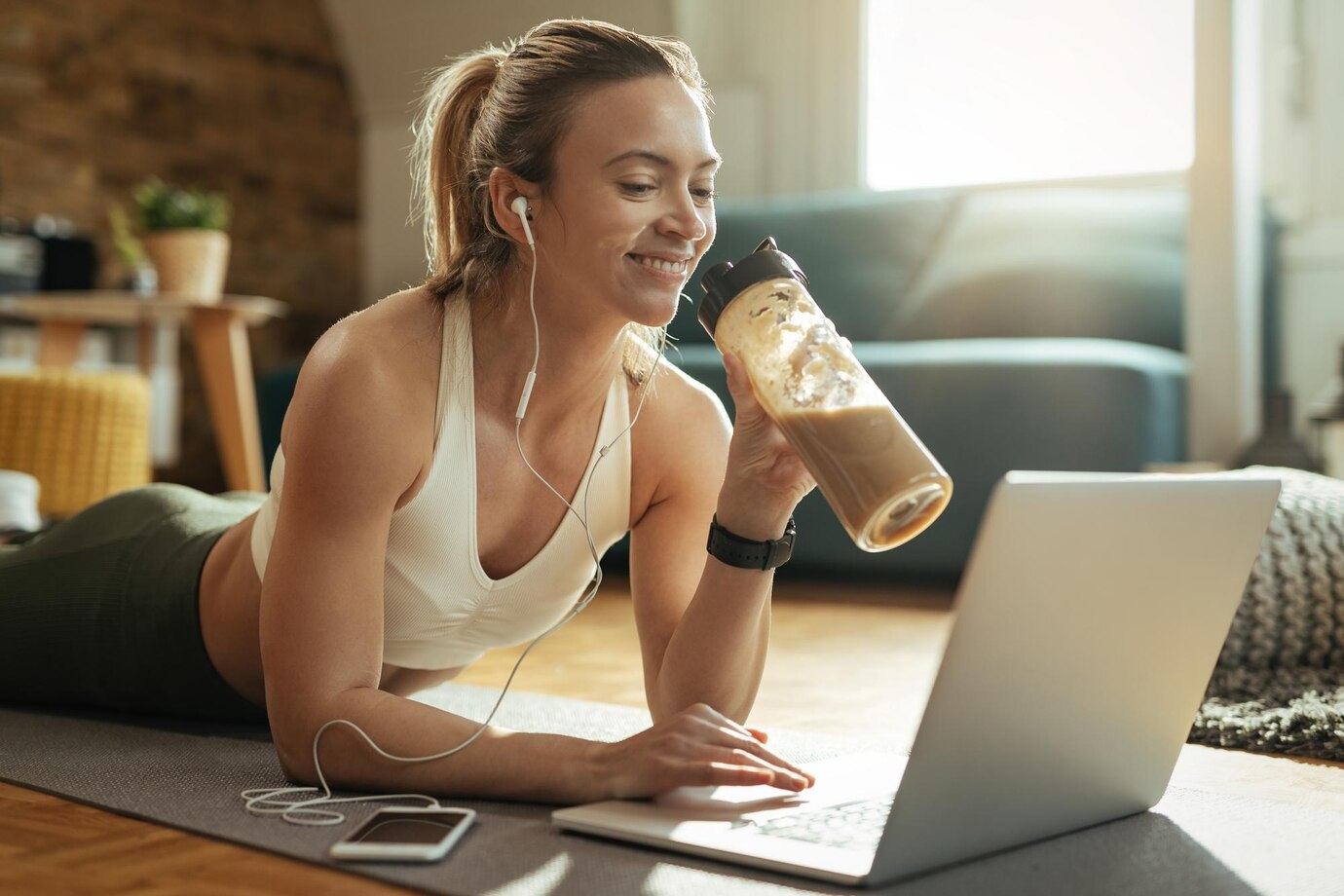 young-happy-sportswoman-drinking-smoothie-while-relaxing-floor-surfing-net-laptop_637285-3475