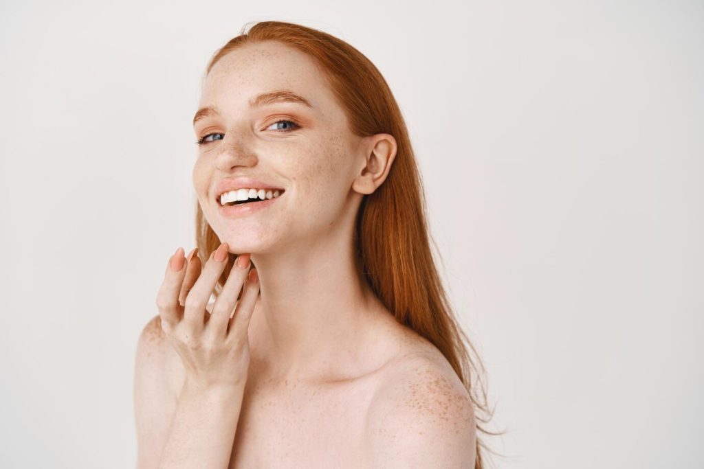 close-up-smiling-redhead-woman-with-pale-skin-freckles-touching-soft-perfect-face-using-skincare-cream-standing-white-wall_176420-42743